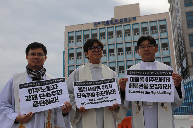 미사를 봉헌한 사제들이&nbsp;부산출입국외국인청을 배경으로 "이주노동자 강제 단속과 강제 추방을 중단하고 체류권을 보장하라"는 손자보를 들고 있다. (왼쪽부터) 쩐국풍(TRAN QUOC PHONG) 노동사목 베트남 전담 신부, 김진호 노동사목 부본부장 신부, 차광준 노동사목 본부장 신부.&nbsp;ⓒ장영식<br>